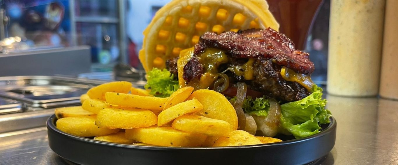 Burger mit Waffelbrötchen, Salat, Bacon und frittierten Bratkartoffeln auf einem schwarzen Teller.